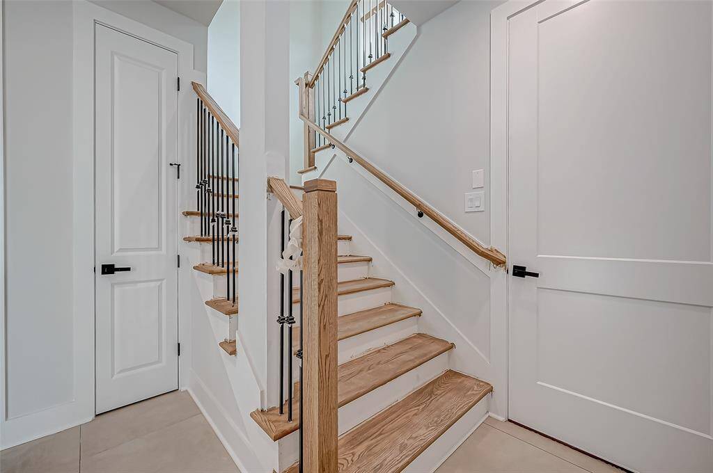 This photo shows a modern entryway with a sleek black front door featuring glass panels, allowing natural light to enter. Adjacent is a white door opening to a storage area under stairs, and a staircase with wooden steps and black metal railings. The space has light-color walls and flooring, creating a bright and welcoming ambiance.