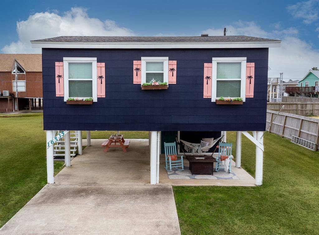 Elevated coastal home with a charming navy exterior and pink shutters. Features a covered patio area with seating and a hammock underneath. Ideal for enjoying outdoor relaxation.