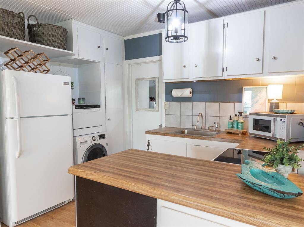 Cozy kitchen with a warm, rustic feel featuring wood countertops and white cabinetry. It includes modern appliances like a refrigerator, washer, and microwave. The space is well-lit with a charming hanging light fixture, and there are decorative touches like baskets and plants for added character.