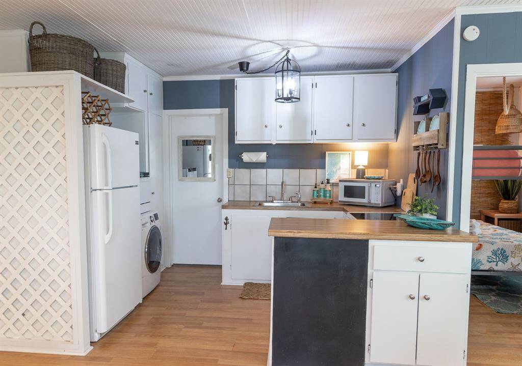 This cozy kitchen features white cabinetry, wooden countertops, and a tile backsplash. It includes essential appliances like a refrigerator, microwave, dishwasher and a washer-dryer combo. A small island offers additional counter space, while open shelving provides storage.