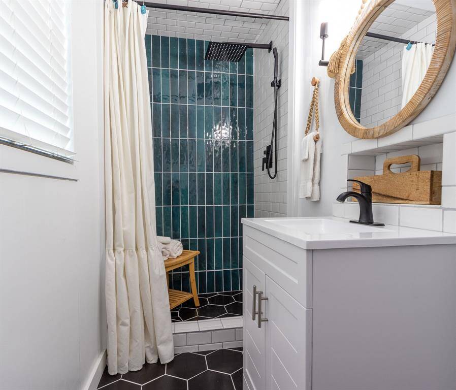 This bathroom features a modern design with a walk-in shower showcasing striking teal tiles. It includes a black rainfall showerhead, hexagonal floor tiles, and a clean, white vanity with a round mirror. Natural light filters in through a window with blinds.