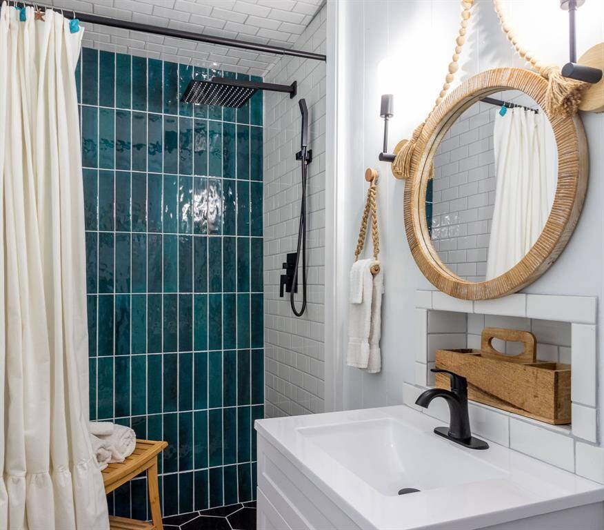 This bathroom features a modern shower with teal tiles and a rainfall showerhead creating a zen/spa like feel.  It has a sleek black faucet, a round woven mirror, and a white vanity. The decor is minimalist with a touch of natural elements.  A water heater installed Jan 2025.