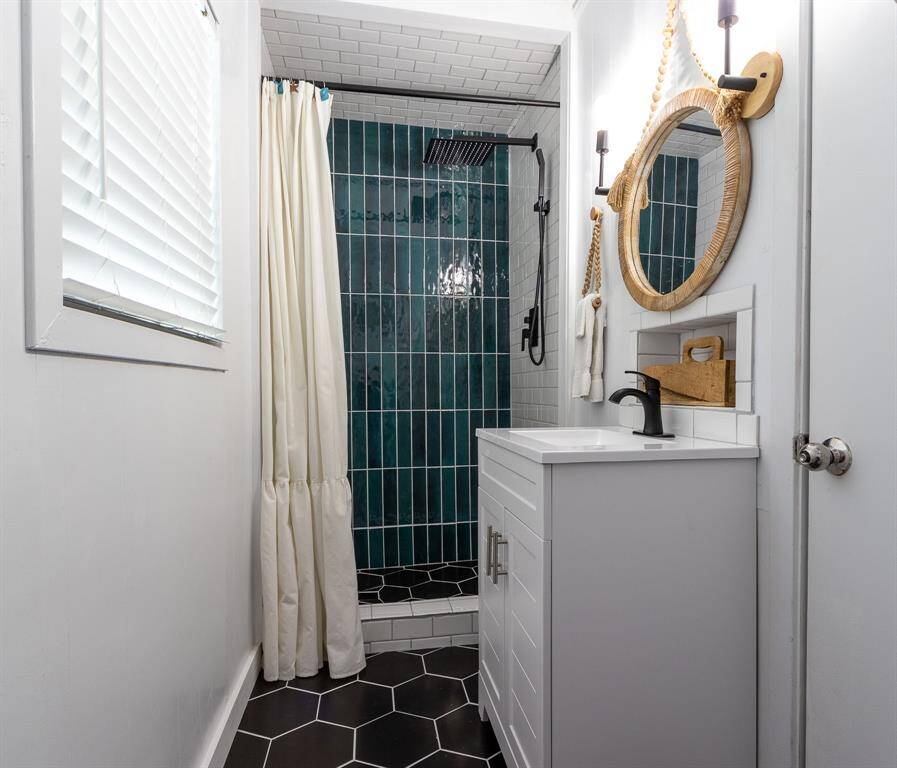 This bathroom features a modern design with a sleek black hexagonal tile floor and a striking teal tiled shower. It includes a rainfall showerhead with a spray handle, a white vanity with a round mirror accented by a rope frame, and contemporary fixtures, creating a stylish and functional space.