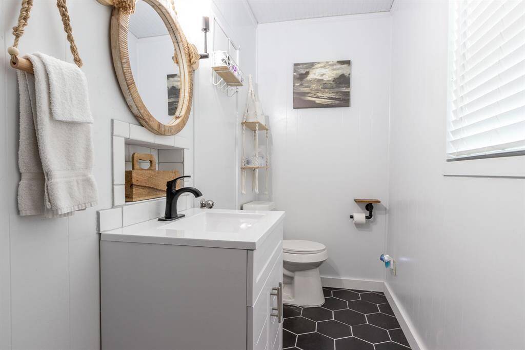 A modern bathroom with a sleek white vanity and black faucet. The space features hexagonal floor tiles, a round mirror with a rope accent, and minimalist decor. Natural light filters through a window with blinds, adding brightness to the room.
