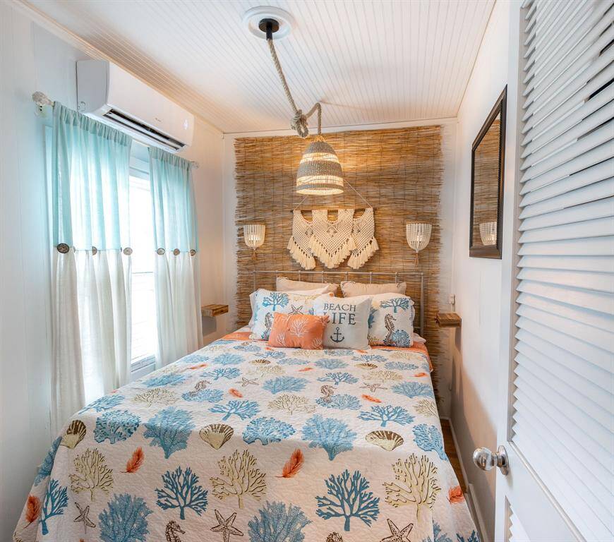 Cozy bedroom with a beachy theme, featuring nautical bedding, and soft blue curtains. It includes a pendant light and mini split air conditioning for comfort.