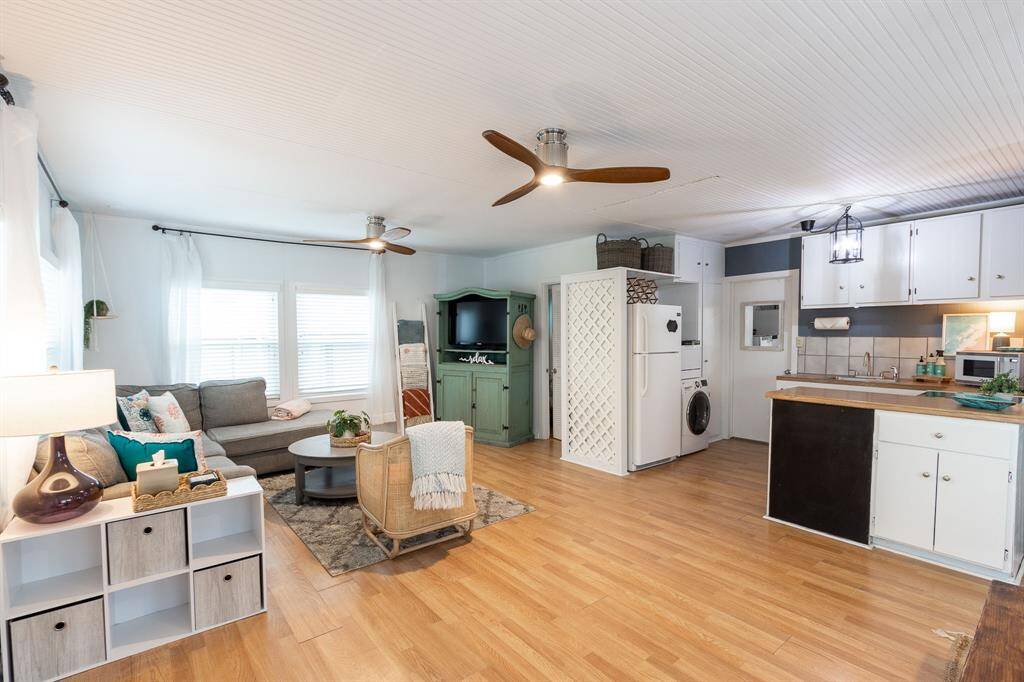 A light/bright, open-concept living area with laminate floors, a cozy sectional sofa, and a ceiling fan. The space features a blend of modern and rustic elements, including a kitchen with white cabinetry, and ample natural light from large hurricane impact windows.