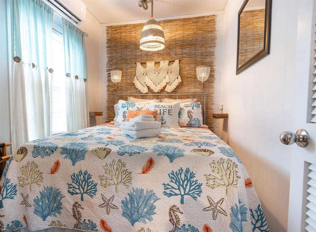 This cozy bedroom features a coastal theme with a textured woven accent wall, a bed adorned with seashell and coral-patterned bedding, and soft natural lighting through light blue curtains. Perfect for a serene beach-style retreat.