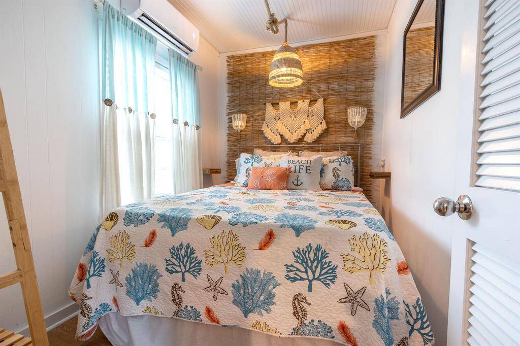 This cozy bedroom features a beach-themed decor with a colorful coral-patterned bedspread and decorative pillows. Light blue curtains allow natural light to brighten the space, complemented by a woven headboard and pendant light for a coastal vibe.