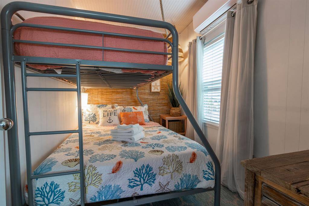 This cozy bedroom features a bunk bed with a nautical-themed comforter, perfect for a beach-inspired decor. The room is well-lit with natural light from the window and has a small wooden nightstand.