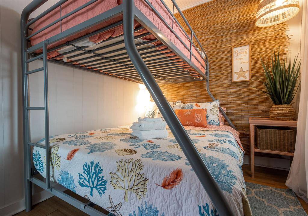This cozy bedroom features a modern bunk bed with a beach-themed quilt on the lower bed. The room has a relaxed coastal vibe with a bamboo accent wall, a wicker lamp, and matching decor. Perfect for a guest or kids' room.
