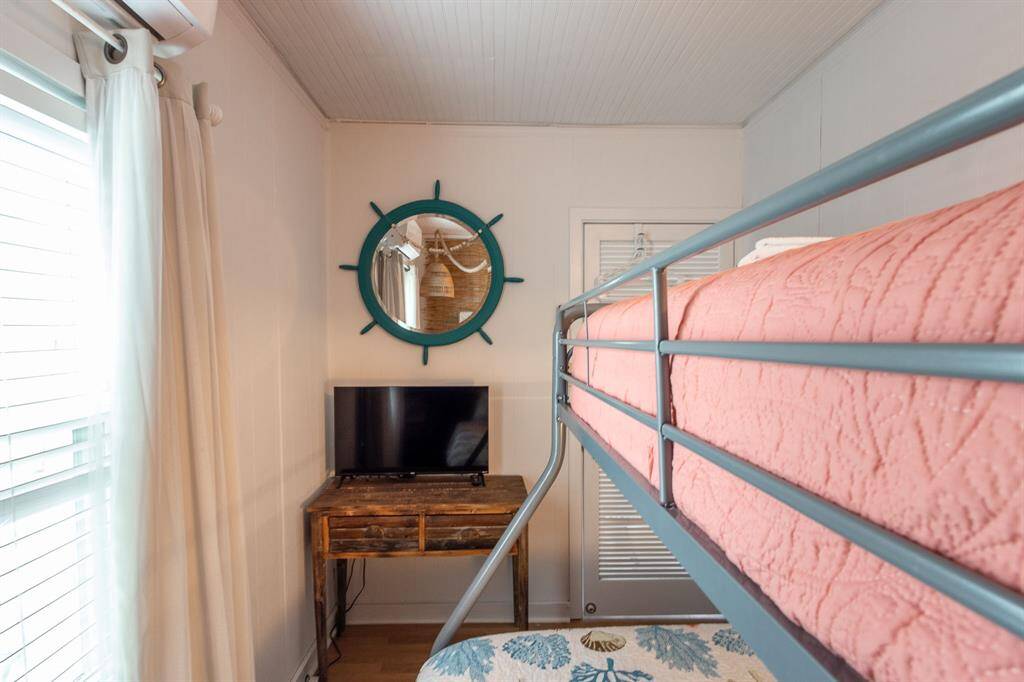 Cozy bedroom with a bunk bed, small TV on a rustic desk, and a nautical-themed mirror. Bright, natural light from a window with white curtains enhances the space.