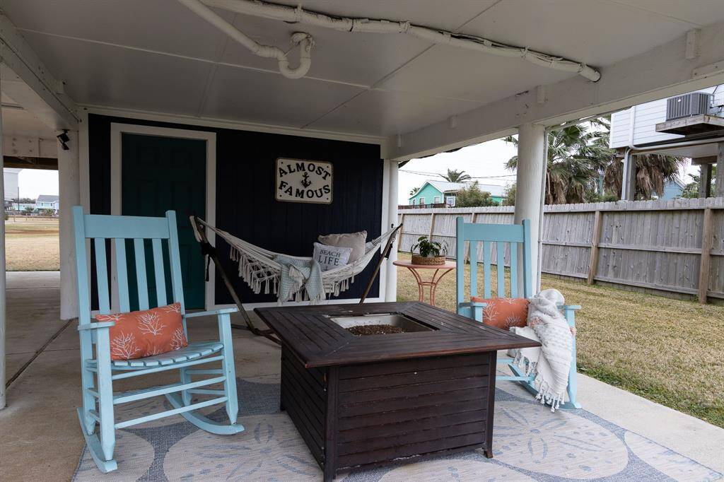 Charming outdoor covered patio with light turquoise rocking chairs, a central gas fire pit, hammock, and decorative touches, perfect for relaxation and entertaining.