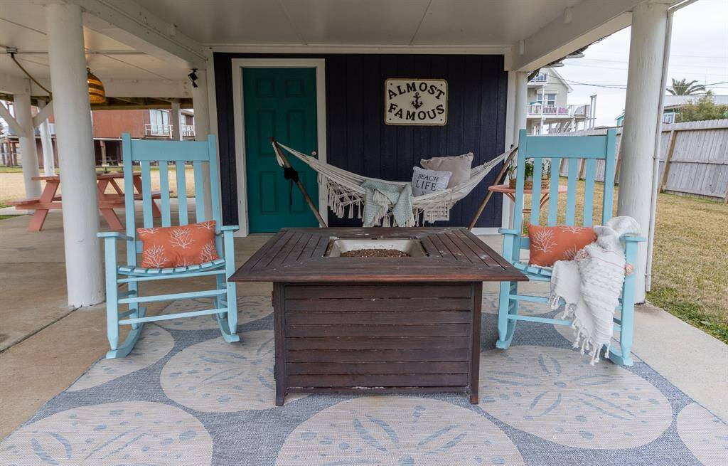 Cozy outdoor seating area with turquoise rocking chairs, a hammock, and a gas fire pit table on a decorative rug. The space is inviting and perfect for relaxation.