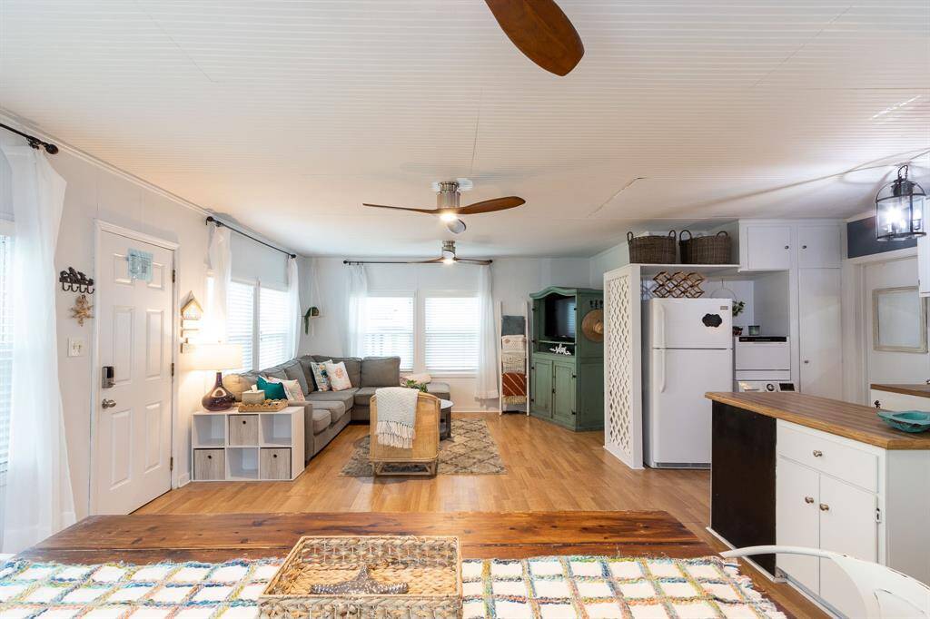 This bright and cozy open-concept living space features a modern kitchen with white cabinetry and a laminate butcher block countertop, a comfortable seating area with a sectional sofa, and stylish ceiling fans. Natural light floods the room through large windows, creating an inviting atmosphere.