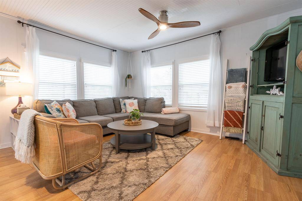This cozy living room features a large sectional sofa, a stylish ceiling fan, and ample natural light from multiple windows. The space is accented with a soft area rug, a round coffee table, and a charming green cabinet for entertainment needs.