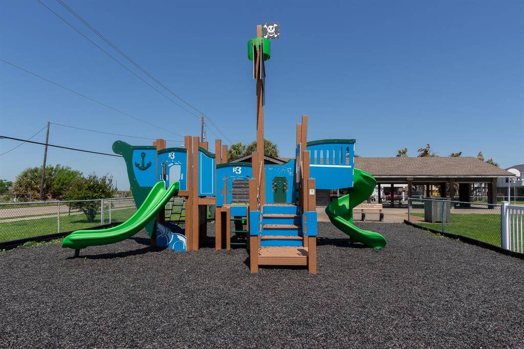 This photo showcases a vibrant, pirate-themed playground with slides and climbing features, set on a safe rubber surface. It's an inviting space for children to play, located near a residential area with a fenced yard and a pavilion in the background just steps away.