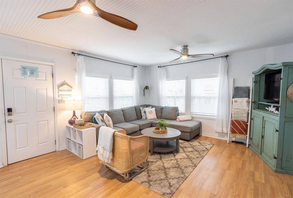 This cozy living room features a light, airy design with a large sectional sofa, laminate wooden floors, and modern ceiling fans. Natural light pours in through multiple windows, highlighting the stylish decor and a charming entertainment unit. Perfect for relaxed living.