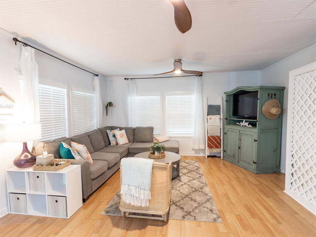 This cozy living room features a large sectional sofa, a rustic green entertainment cabinet, and bright natural light from multiple windows. The laminate wooden floor and ceiling fan add warmth and comfort, while a stylish area rug and decor complete the inviting space.