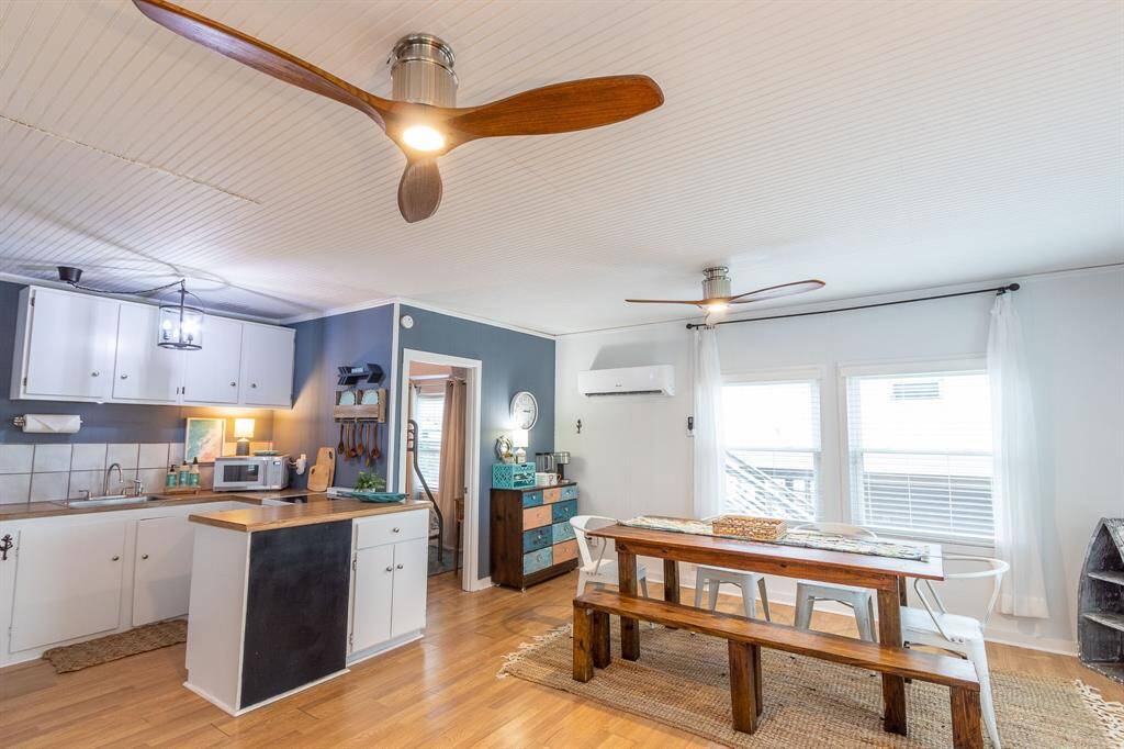 Bright, open-concept kitchen and dining area. The kitchen features white cabinetry, a tiled backsplash, and modern appliances. The dining space includes a rustic wooden table with bench seating. Ceiling fans and large windows enhance the airy feel.