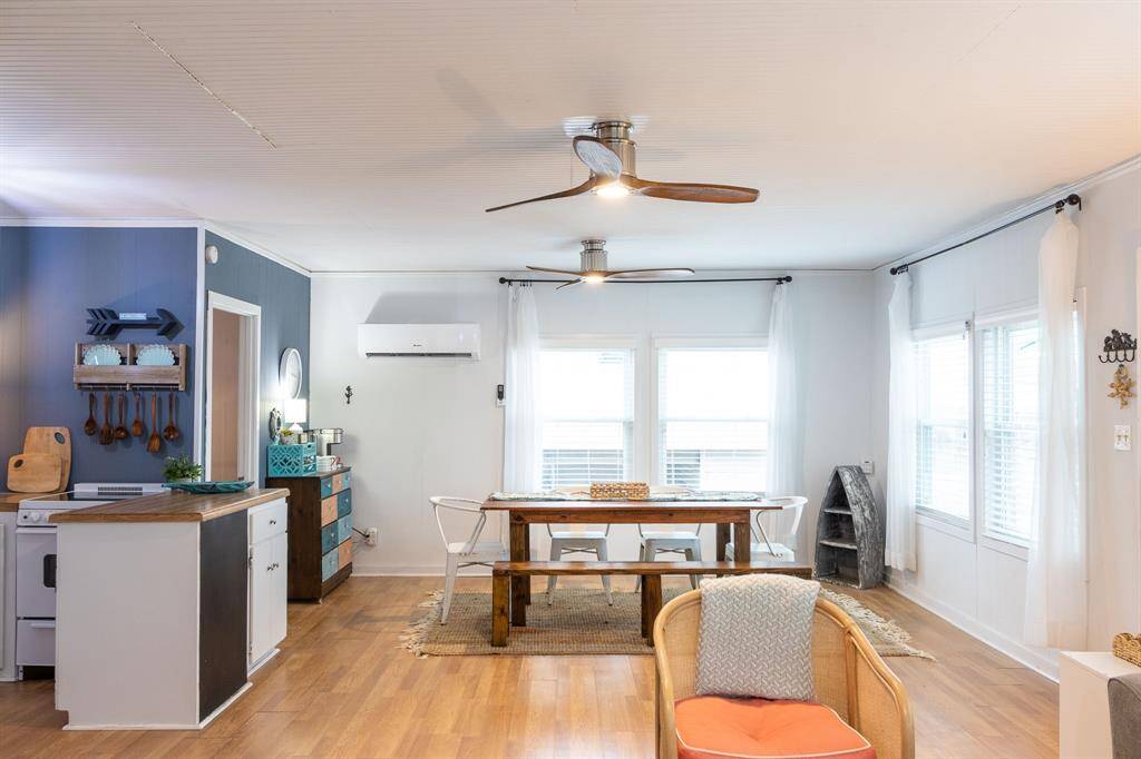 This inviting open-concept living space features warm laminate flooring, a cozy dining area with natural light from large windows, and a modern ceiling fan. The kitchen is compact with stylish cabinetry and a functional island. The room's neutral tones create a bright and airy atmosphere, perfect for comfortable living.