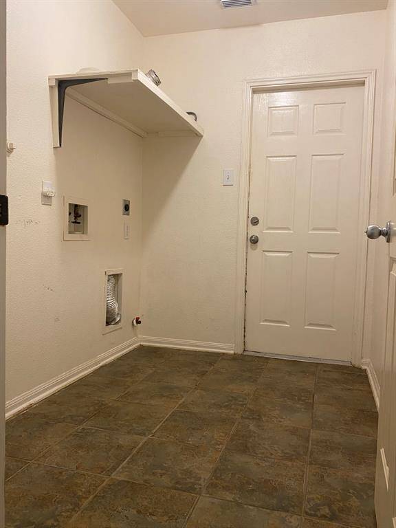 This photo shows a utility or laundry room with tiled flooring, a wall-mounted shelf, and hookups for a washer and dryer. There's a white door leading outside or to another room.