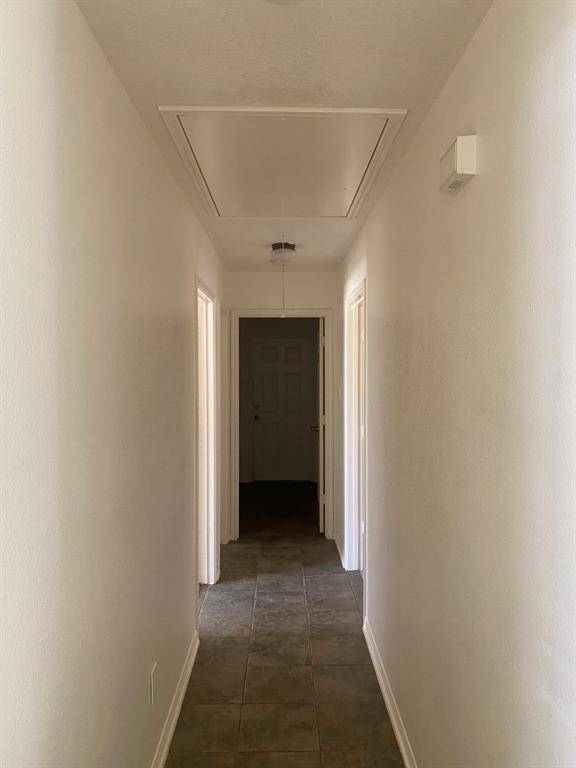 This photo shows a hallway with tiled flooring, white walls, and a ceiling hatch. The hallway leads to a door at the far end and has doorways on both sides.