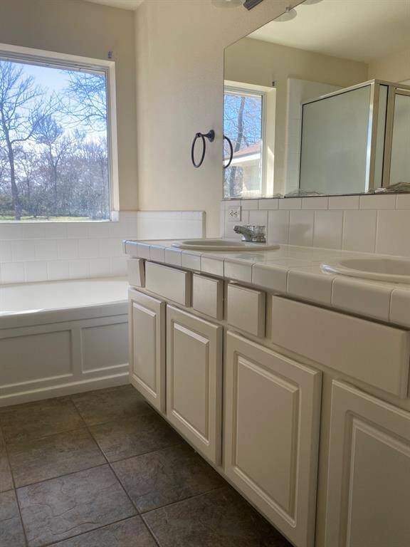 This bathroom features a large window with natural light, a dual-sink vanity with white cabinetry, a tiled countertop, and a spacious bathtub with a separate glass-enclosed shower.