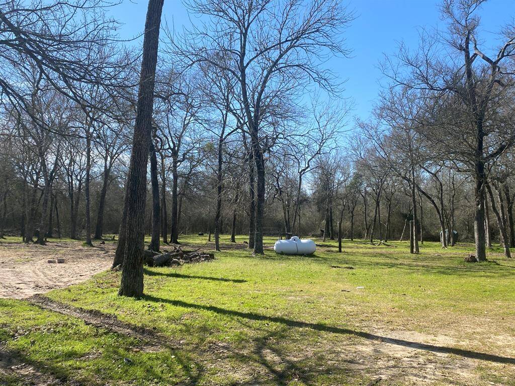 This photo shows a spacious, open yard with scattered trees and a grassy area. There are visible propane tanks, suggesting utility access. The setting is rural and peaceful, ideal for someone seeking a natural environment.