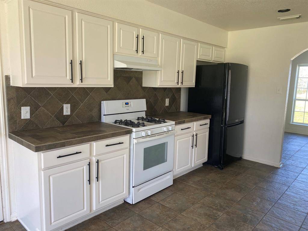 This kitchen features white cabinetry with dark hardware, a gas stove, a black refrigerator, and a tile backsplash. The space has a clean, modern feel with tiled flooring and a view into a bright adjoining room.