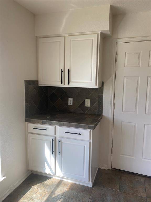 This photo shows a small kitchen area with white cabinets and black handles. The countertop and backsplash feature dark, textured tiles. There is a white door next to the cabinets, and the floor is tiled in a similar style to the backsplash.