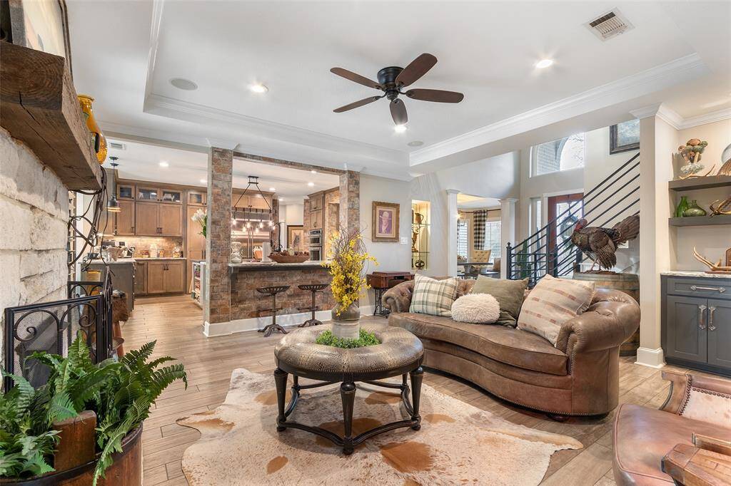 This inviting living area features an open-concept design with a cozy stone fireplace, rich wood flooring, and a seamless flow into a modern kitchen with brick accents. The room is well-lit with natural light, complemented by crown molding, lighted art niche, and a ceiling fan for comfort.