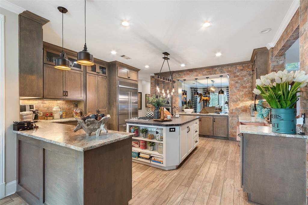This kitchen features a warm, rustic design with wood cabinetry, a central island, and modern stainless steel appliances. The brick accent wall adds character, while ample counter space and lighting create a welcoming, functional space.