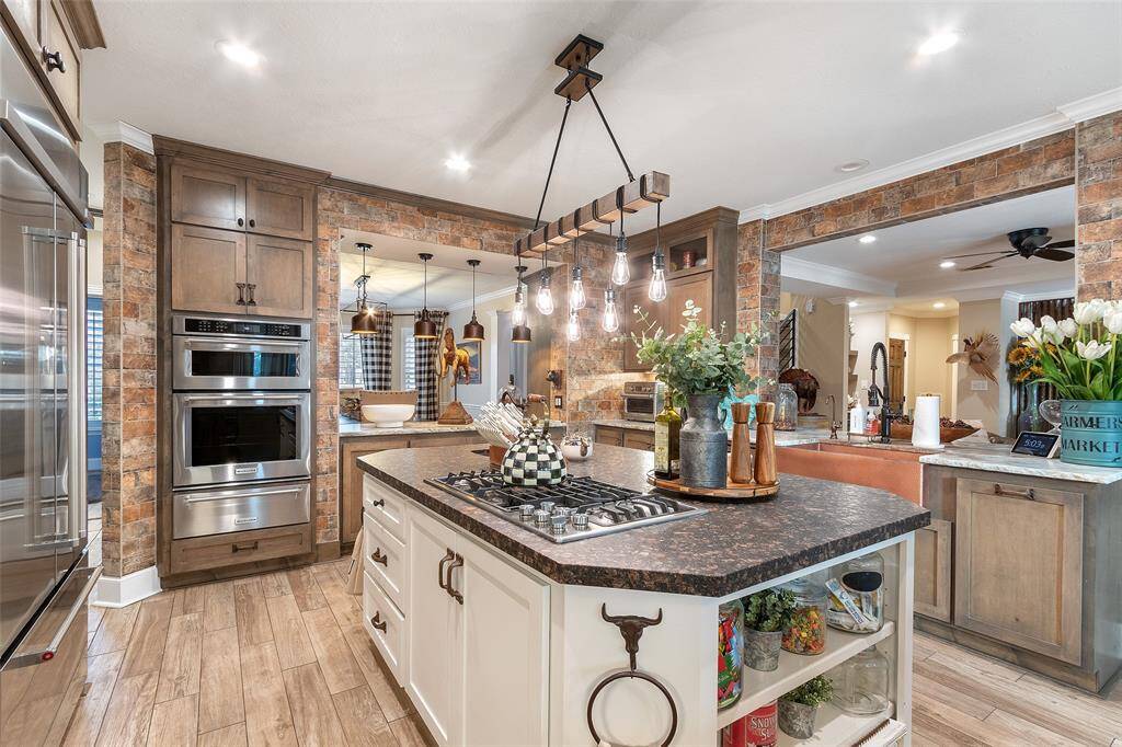 This photo showcases a modern kitchen with a warm, rustic charm. It features a spacious island with a built-in gas cooktop and open shelving. The kitchen is adorned with pendant lighting and brick accents, offering ample cabinetry and stainless steel appliances. The open layout connects seamlessly to the surrounding living areas, ideal for entertaining.