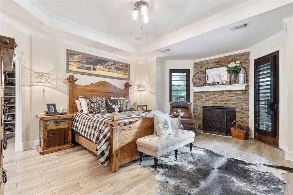This cozy primary bedroom features a stone fireplace, creating a warm, inviting atmosphere. It has a stylish tray ceiling, elegant lighting, crown molding, wood floors that enhances the comfort and charm of the space.