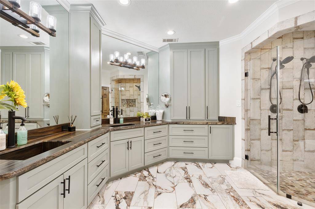 This bathroom features elegant cabinetry with ample storage, a gorgeous granite countertop, and a stylishly tiled walk-in shower. The lighting is modern, complementing the neutral tones and creating a luxurious atmosphere.