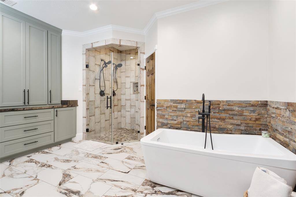 This bathroom features a spacious design with a modern freestanding bathtub, stone accent walls, and a glass-enclosed shower. The room includes elegant cabinetry and a marble-patterned floor, creating a luxurious and contemporary feel.