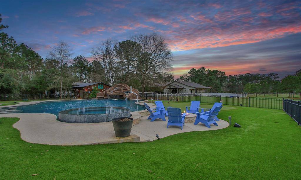 This photo showcases a spacious backyard featuring a large pool with a slide, rock waterfall, a hot tub, and a cozy fire pit area. This beautiful unset sky offer a serene and inviting outdoor living space.