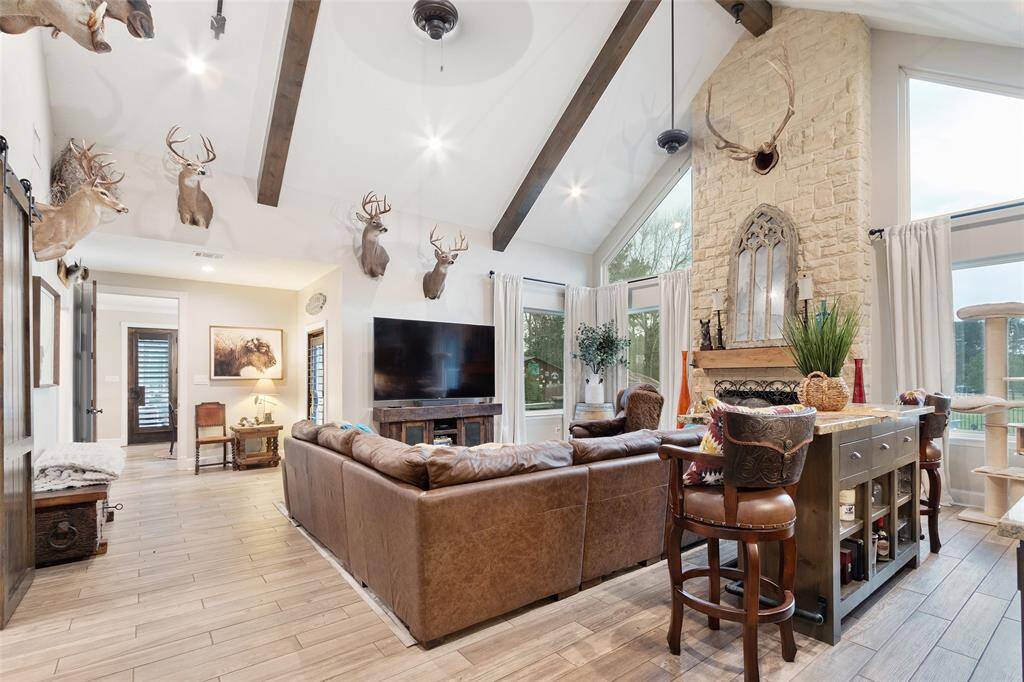 This spacious living room/game room features a high vaulted ceiling with rustic wooden beams, a stone fireplace, and ceiling fans. It has large windows for natural light, perfect for a homey, lodge-like atmosphere.