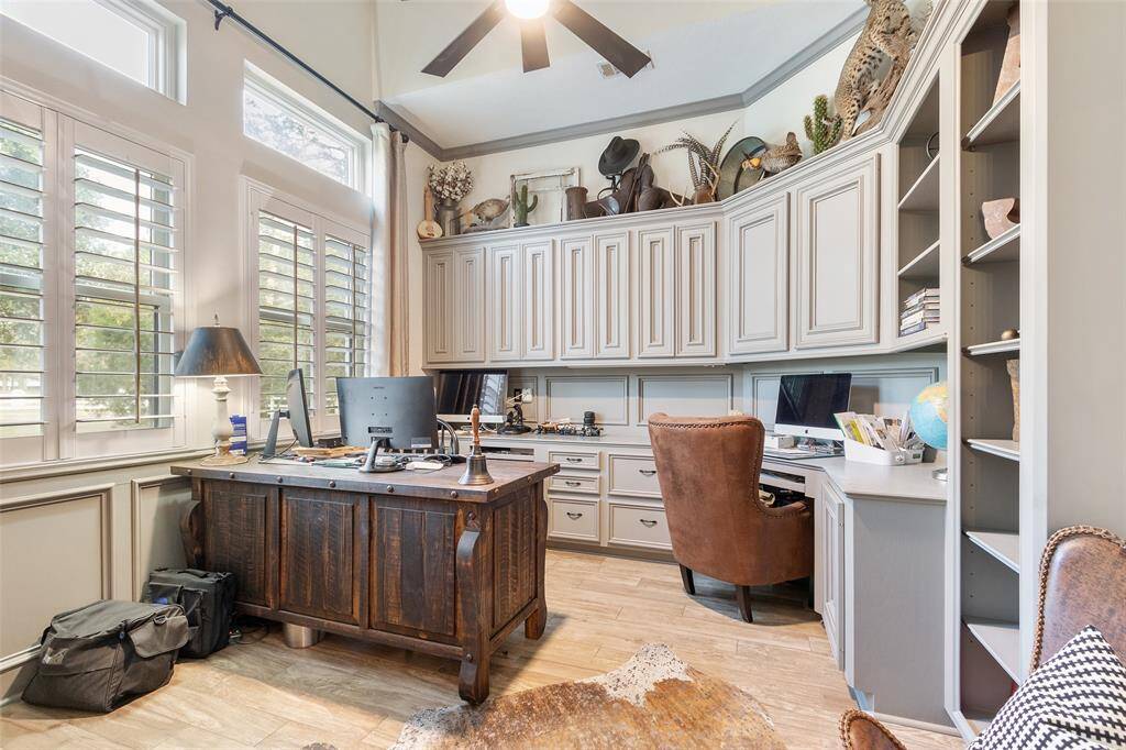 This photo showcases a spacious home office with high ceilings, abundant natural light from large windows, and elegant plantation shutters. The room features built-in shelves and cabinets, creating a cozy yet functional workspace.
