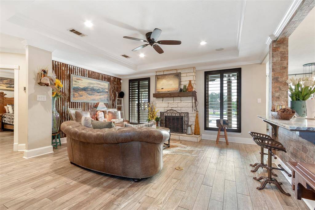 This cozy family room features a rustic stone fireplace, wood flooring, and large windows with shutters.  A stylish open layout that connects to the kitchen area, offering a warm and inviting atmosphere.