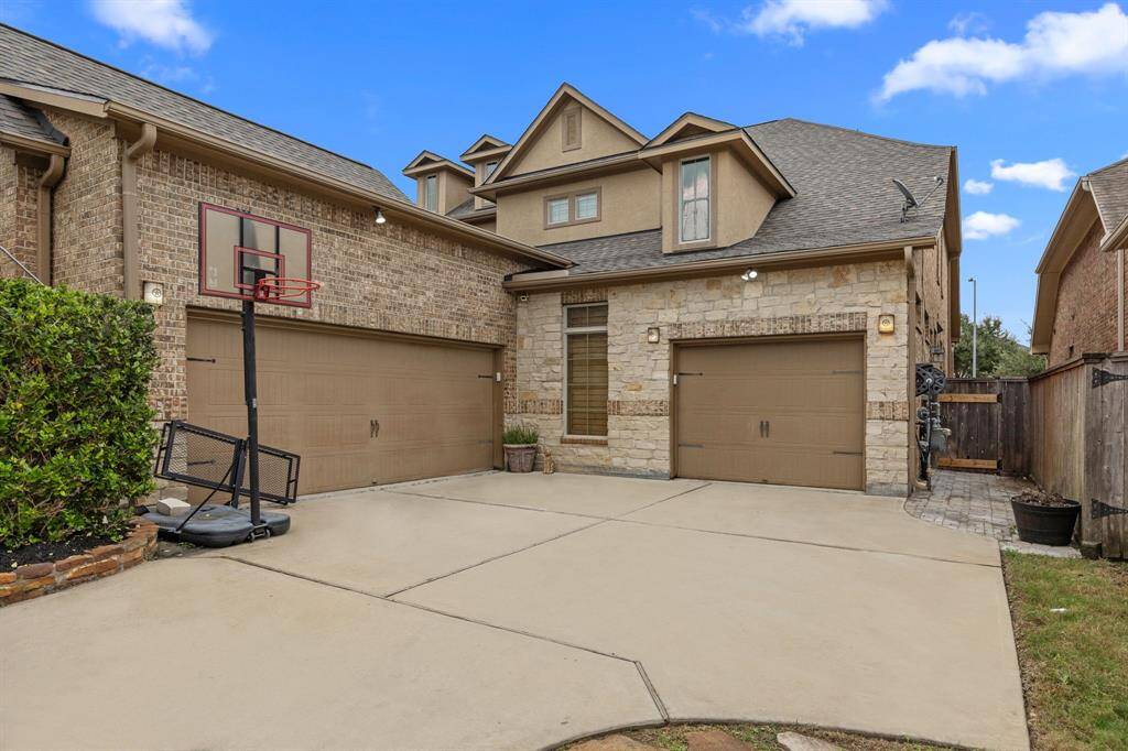 This photo showcases a spacious, two-story brick and stone home with a three-car garage. The driveway includes a basketball hoop, and there's a side walkway leading to the backyard. The exterior features a blend of traditional and modern architectural elements.