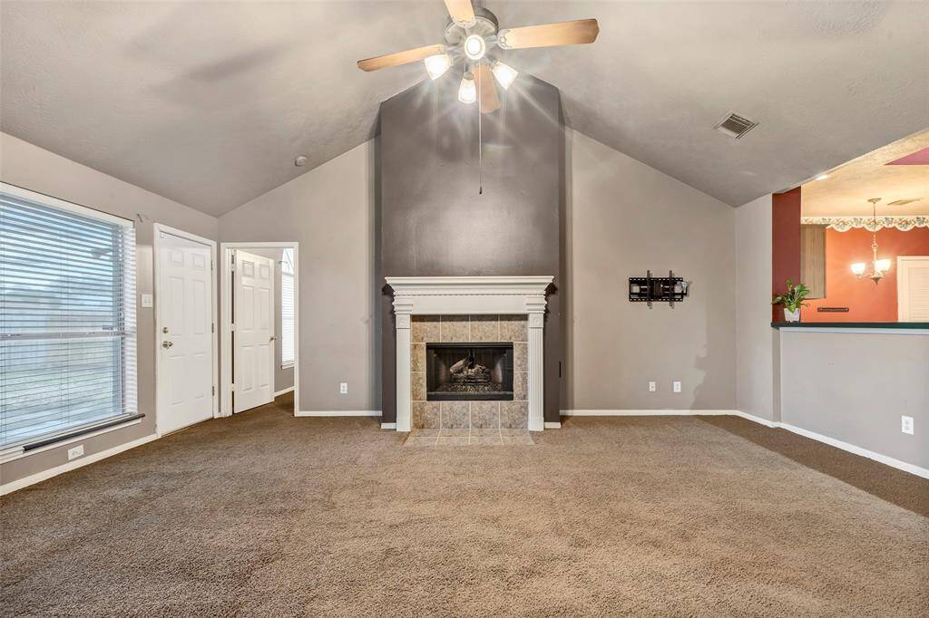 This spacious living room features a high vaulted ceiling with a fan, a cozy fireplace with a stylish mantel, and large windows for ample natural light. The neutral carpet and wall colors create a warm atmosphere, and there's a convenient pass-through to the adjoining kitchen area.