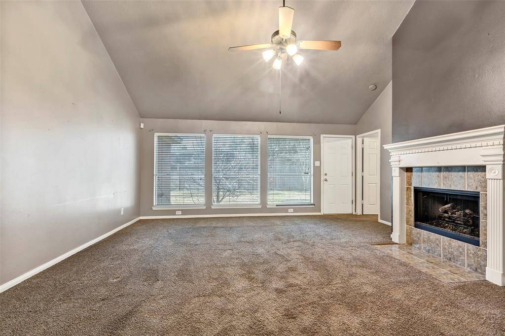 A spacious living room with high ceilings, featuring a ceiling fan and a cozy fireplace. Large windows provide natural light.