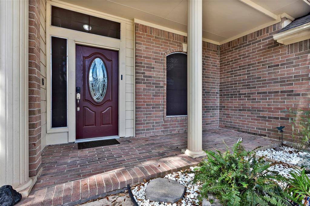 welcoming front porch with a brick exterior, featuring a stylish oval glass front door. The covered entryway is supported by elegant columns, and the area is accented with greenery and white pebble landscaping.