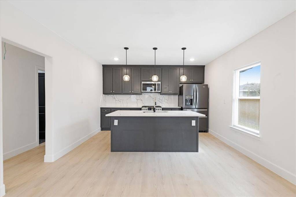 This kitchen features modern dark cabinetry, a central island with pendant lighting, and stainless steel appliances. The light wood flooring and ample natural light create an inviting and spacious atmosphere.