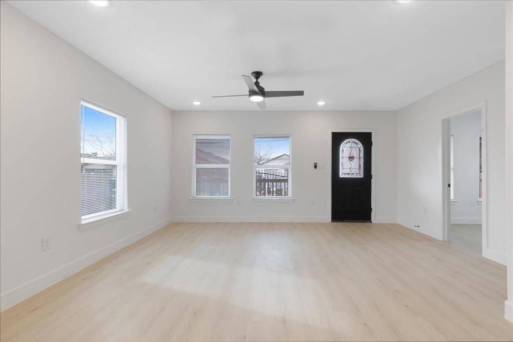 This bright living room features light wood flooring, neutral walls, and ample natural light through multiple windows. It includes a ceiling fan and a stylish black front door, creating a welcoming and modern ambiance.