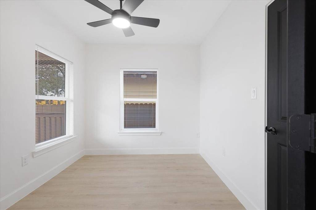 This photo shows a small, bright room with light-colored hardwood flooring and white walls. It features two windows providing natural light, a ceiling fan with a light fixture, and a black door. Perfect for a cozy office or guest room.