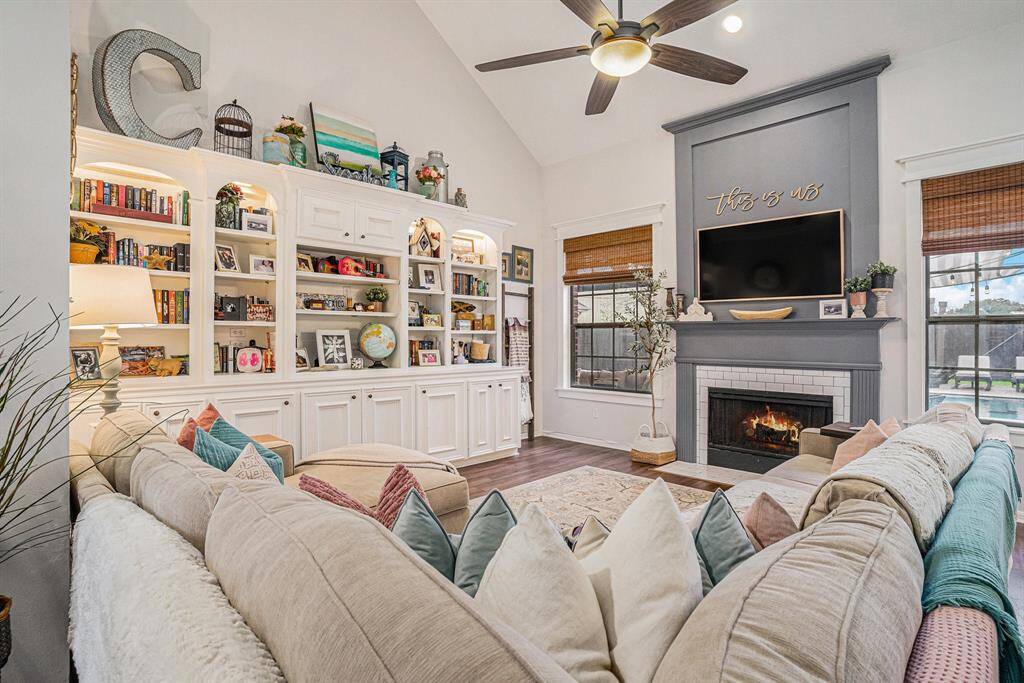 This inviting living room features a cozy fireplace, built-in bookshelves, and a high ceiling with a modern fan. Natural light streams through large picture windows with wooden blinds.