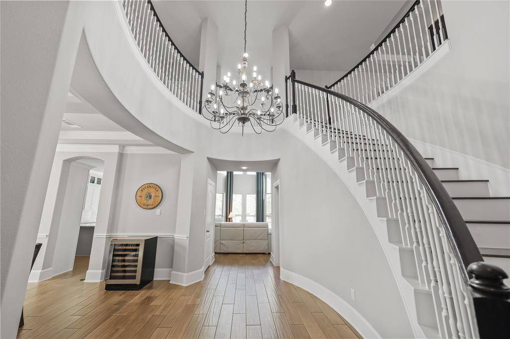 This grand foyer showcases a striking cascading staircase with a dark handrail and white balusters. The soaring ceilings enhance the open and airy feel, while an elegant chandelier serves as a stunning centerpiece, adding a touch of sophistication. The wood-look tile flooring extends throughout the space while the decorative wainscoting and crown molding enhance the architectural appeal.