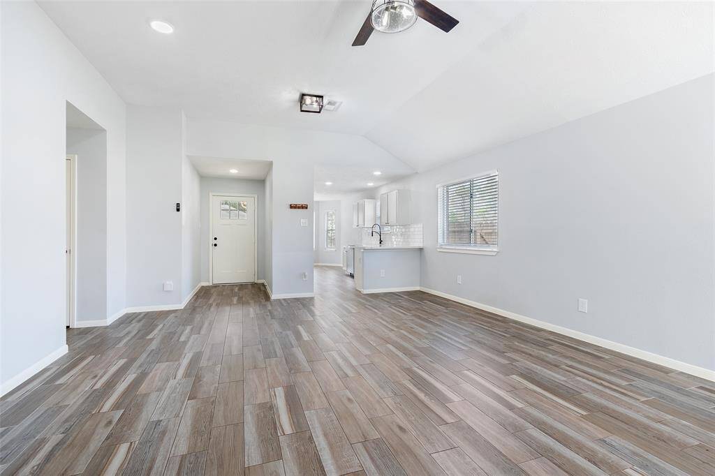 This photo shows a bright, open-concept living space with modern wood-look tile flooring. The room features recessed lighting, a ceiling fan, and flows seamlessly into a contemporary kitchen with white cabinetry and a large window for natural light.
